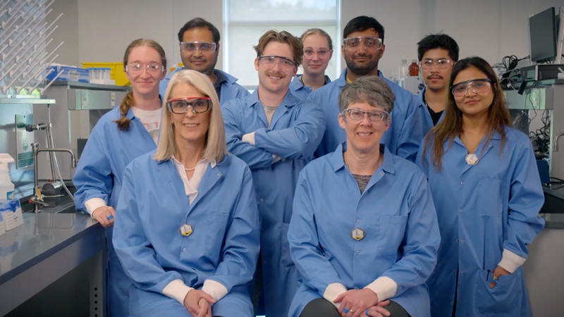 A group of researchers wearing protective glasses and lab coats pose for a portrait in between research benches filled with glassware and equipment