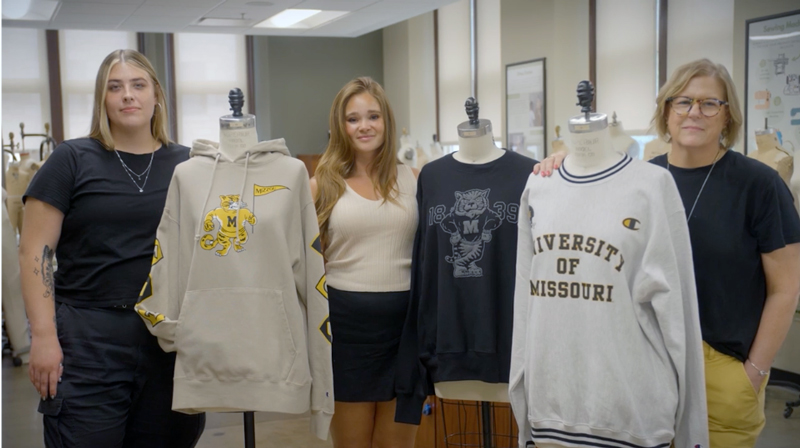 Kerry McBee-Black poses with two other students in a sewing workroom between dress forms covered in student-designed Mizzou sweatshirts