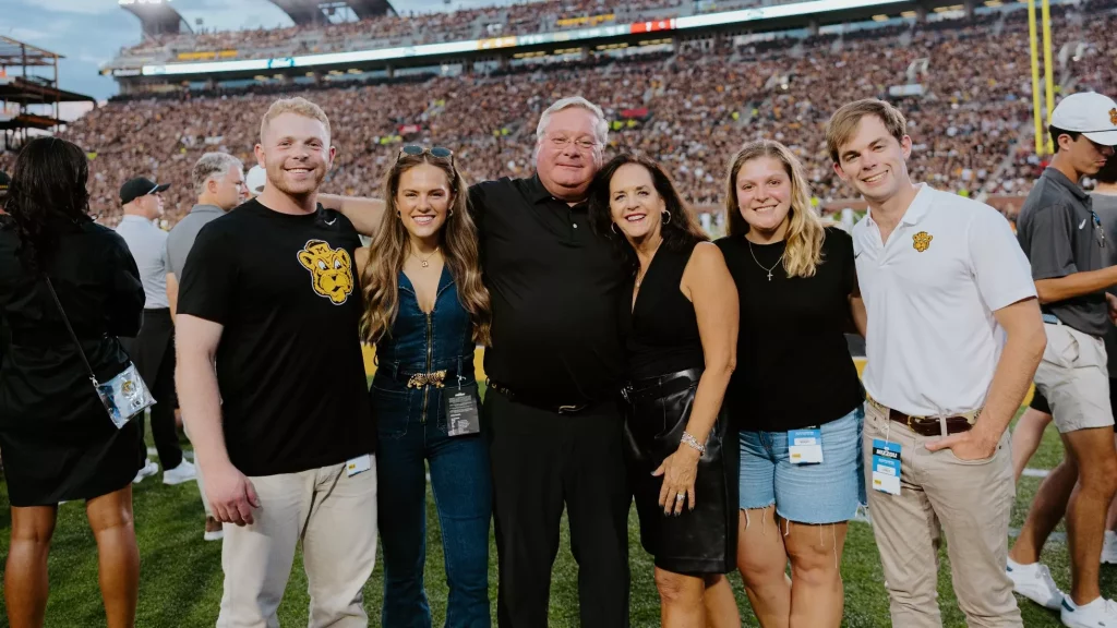 Paul and Lynn Vogel stand with their family outside the end zone of Faurot Field with a packed stadium behind them.