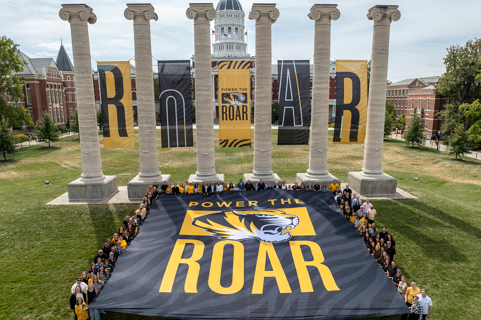 A group of people holding a Power the Roar flag in front of the Columns